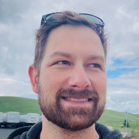 Joe Gilmore smiling outdoors, with green hills and a cloudy sky behind him